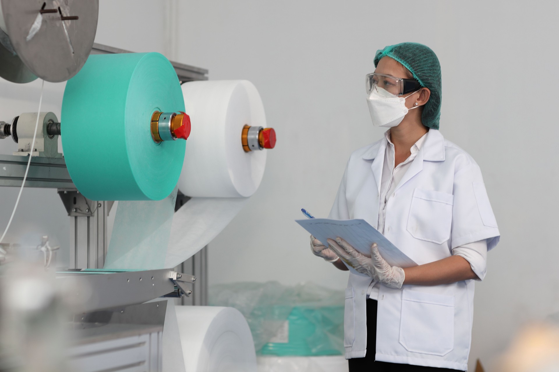 Worker woman in personal protective equipment or PPE inspecting quality of mask and medical face mask production line in factory, manufacturing industry and factory concept.