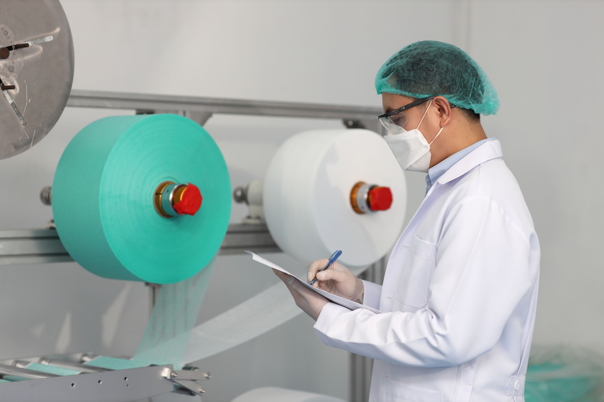 Worker man in personal protective equipment or PPE inspecting quality of mask and medical face mask production line in factory, manufacturing industry and factory concept.